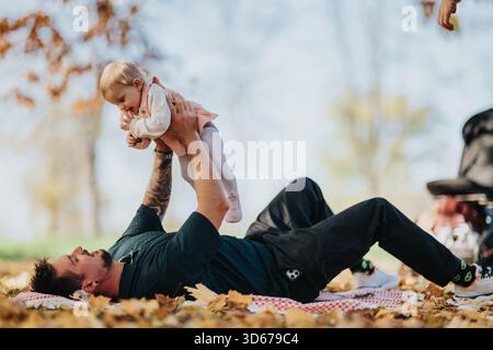 Padre sdraiato sulla coperta nel parco autunnale sollevando la bambina e sorridendo al momento Foto Stock