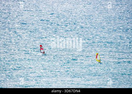 Un teleobiettivo che cattura due windsurfer con vele dai colori vivaci (rosso e giallo/verde) navigando sull'acqua blu instabile e soleggiata al largo del co Foto Stock