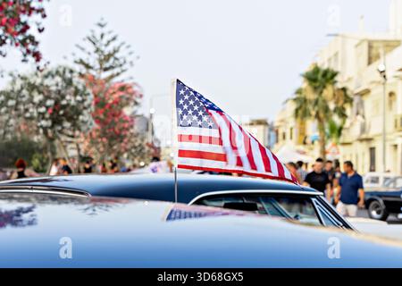 Piccola bandiera americana sul tetto dell'auto in occasione dell'evento cittadino, gente e palme in un soffice sfondo, brillante atmosfera estiva Foto Stock
