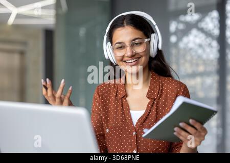 Giovane donna indiana che indossa cuffie e occhiali. Guardare lo schermo di un notebook mentre si tiene in mano un notebook e si gestiscono con una mano. Partecipare attivamente a una riunione virtuale o a una sessione di apprendimento online Foto Stock