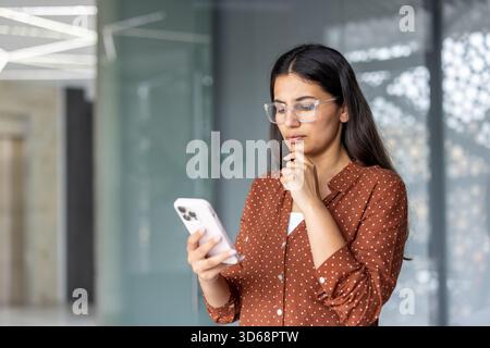 Donna indiana che indossa occhiali e una camicia a pois con uno smartphone, guarda lo schermo con un'espressione premurosa mentre contempla i contenuti digitali in un ufficio moderno Foto Stock
