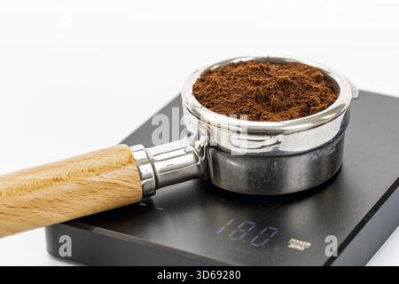 Primo piano di un portafiltro in metallo riempito con caffè espresso appena macinato, appoggiato su una bilancia digitale che mostra una misurazione del peso, con un han di legno Foto Stock