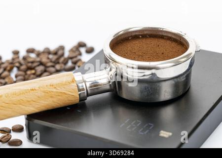 Un portafiltro riempito di caffè finemente macinato si trova su una bilancia digitale che mostra 118,0 grammi, con chicchi di caffè tostati sparsi nel dorso Foto Stock