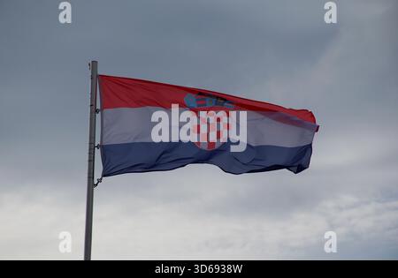 Vibrante bandiera croata con stemma che ondeggia in forte vento sotto un cielo nuvoloso. Patriottismo, identità nazionale, Europa, viaggi. Foto Stock