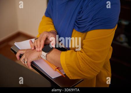 Una donna con maniche blu hijab e senape tiene con sé documenti e un notebook mentre controlla uno smartwatch in un ufficio contemporaneo, ritraendo gli impegni quotidiani Foto Stock