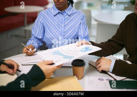 Gruppo multietnico di giovani professionisti adulti che collaborano a tavola, analizzando grafici e grafici finanziari, scambiando documenti, discutendo strategia aziendale durante la riunione Foto Stock