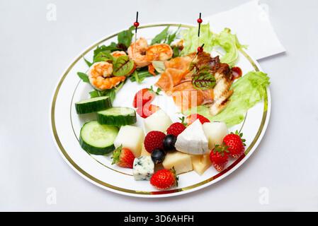Un piatto gourmet splendidamente organizzato con frutta vivace, succulenti gamberi, salmone affumicato e una selezione di formaggi su un piatto bianco. perfetto Foto Stock