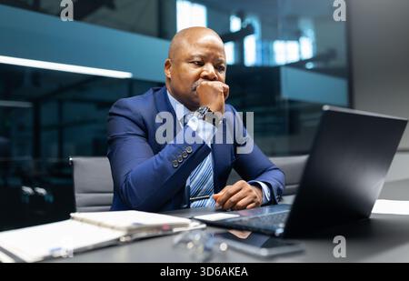 Uomo d'affari nero maturo seduto alla scrivania, pensando seriamente mentre guarda un notebook Foto Stock