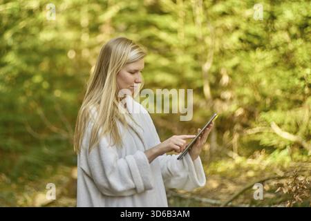 Donna in accappatoio con tablet in mezzo alla natura, benessere, relax, spa resort, giardino, esterni, connettività, digitale, online, internet, riposiziona arco Foto Stock