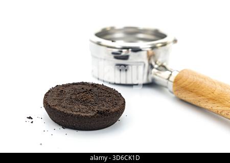 Immagine ravvicinata dei fondi di caffè compattati usati posizionati su una superficie bianca accanto a un portafiltro metallico con manico in legno, evidenziando la presenza di caffè Foto Stock
