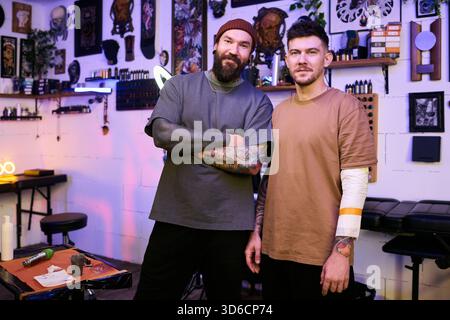 Due giovani adulti caucasici in piedi in studio di tatuaggio, entrambi con braccia tatuate, un braccio incrociato e sorridenti, altri con braccio in bendatura, attrezzature per tatuaggi visibili sullo sfondo Foto Stock
