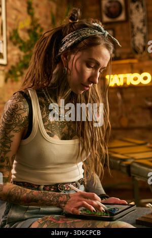 Giovane donna caucasica adulta con dreadlock lunghi e tatuaggi estesi seduta in casa con smartphone, concentrandosi sullo schermo, indossando gioielli e fascia, sfondo visibile dello studio di tatuaggio Foto Stock