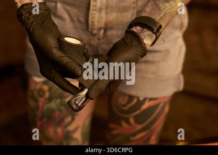 Giovane donna caucasica tatuata che indossa guanti neri per preparare la macchina per il tatuaggio con bendaggio, in piedi in studio con tatuaggi dettagliati sulle gambe visibili Foto Stock