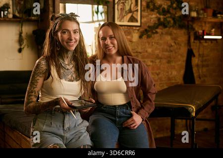 Ritratto di due giovani donne caucasiche sedute insieme nello studio del tatuaggio, una donna pesantemente tatuata che tiene un tablet digitale e sorride, entrambe guardando la fotocamera Foto Stock