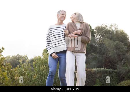 Due donne in maglione a righe e cardigan marrone che camminano braccio in braccio nel verde del parco, sorridenti Foto Stock