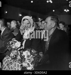 Slavyansk, Ucraina, URSS - anni '1970: Ritratto d'archivio emotivo di una coppia anziana in udienza al Palazzo Artem della Cultura. Una donna con il velo si strappa le lacrime dagli occhi tenendo in mano un mazzo di fiori. Indossa medaglie sulla sua giacca. L'uomo accanto a lei guarda seriamente il palco. Questa immagine in bianco e nero cattura un momento toccante durante un concerto festivo o una cerimonia d'onore nella regione di Donetsk, riflettendo i sentimenti profondi della vecchia generazione nella pacifica era sovietica. Foto Stock