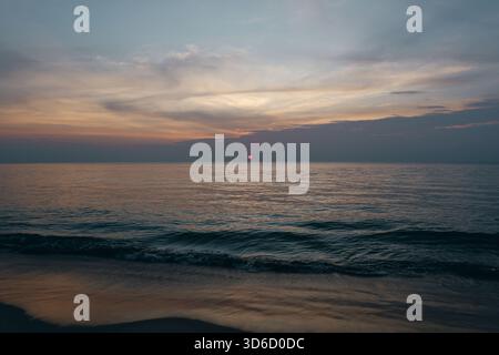 Il sole che tocca l'orizzonte e porta una luce calda alle dolci onde che si lavano sulla spiaggia, catturando un momento di relax e la bellezza panoramica della costa. Foto Stock