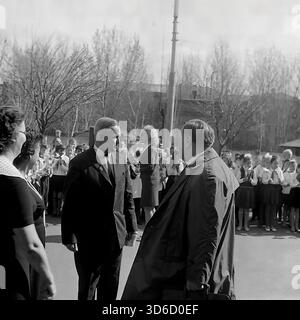 Slavyansk, Ucraina, URSS - 1970s: Fotografia d'archivio di una conversazione tra l'amministrazione scolastica e un ospite ufficiale durante un'assemblea festiva alla Scuola No. 12. Due uomini in tuta formale parlano nel cortile della scuola, mentre le file di studenti con archi bianchi applaudono sullo sfondo. Questa immagine in bianco e nero cattura l'atmosfera di un evento solenne, probabilmente un'induzione dei pionieri o una vacanza, che riflette le tradizioni educative della regione di Donetsk durante la pacifica era sovietica. Foto Stock