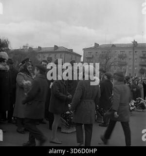 Slavyansk, Ucraina, URSS - 1970: Vivace scena d'archivio in Piazza della Rivoluzione d'ottobre durante la celebrazione del 7 novembre. I bambini delle scuole presentano fiori a funzionari e veterani, mentre un fotografo cattura il momento. I cittadini sono vestiti con cappotti caldi e cappelli per il freddo autunno. Edifici residenziali sovietici sono sullo sfondo. Questa immagine in bianco e nero riflette l'atmosfera festosa, lo spirito della comunità e le tradizioni della regione di Donetsk durante la pacifica era sovietica. Foto Stock