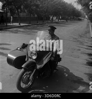 Slavyansk, Ucraina, URSS - 1970: Fotografia d'archivio di un ufficiale della polizia stradale sovietica (GAI) che pattuglia le strade della città su una motocicletta di servizio con un sidecar. Il poliziotto indossa un'uniforme e un berretto mentre guida una moto pesante d'epoca dotata di parabrezza e copertura in tela. Sullo sfondo, un'auto GAZ-21 Volga e un ciclista sono visibili su un tranquillo viale alberato. Questa immagine in bianco e nero cattura l'atmosfera dell'ordine pubblico e della vita quotidiana nella regione di Donetsk durante l'era pacifica. Foto Stock
