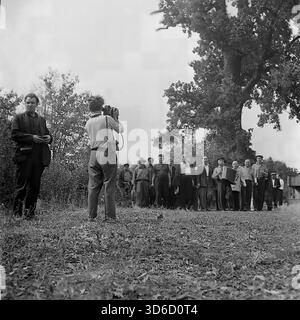 Slavyansk sobborgo, Ucraina, URSS - anni '1970: Fotografia d'archivio dinamica di una processione rurale festosa. Un gruppo di uomini, guidati da un suonatore di fisarmonica, cammina lungo un sentiero vicino a una grande quercia. Un cameraman con una videocamera filma la scena, davanti alla colonna. Questa vivace immagine in bianco e nero cattura l'atmosfera di una vacanza del villaggio (forse un festival del raccolto), le tradizioni popolari e la copertura mediatica nella regione di Donetsk durante la pacifica era sovietica. Foto Stock
