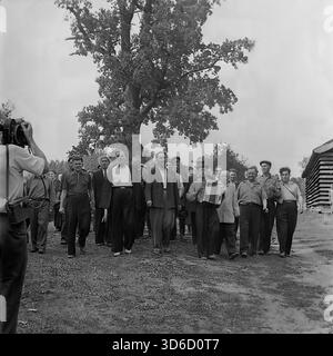 Slavyansk sobborgo, Ucraina, URSS - anni '1970: Fotografia d'archivio dinamica di una processione rurale festosa. Un gruppo di uomini, guidati da un suonatore di fisarmonica, cammina lungo un sentiero vicino a una grande quercia. Un cameraman con una videocamera filma la scena, davanti alla colonna. Questa vivace immagine in bianco e nero cattura l'atmosfera di una vacanza del villaggio (forse un festival del raccolto), le tradizioni popolari e la copertura mediatica nella regione di Donetsk durante la pacifica era sovietica. Foto Stock
