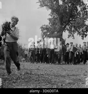 Slavyansk sobborgo, Ucraina, URSS - anni '1970: Fotografia d'archivio dinamica di una processione rurale festosa. Un gruppo di uomini, guidati da un suonatore di fisarmonica, cammina lungo un sentiero vicino a una grande quercia. Un cameraman con una videocamera filma la scena, davanti alla colonna. Questa vivace immagine in bianco e nero cattura l'atmosfera di una vacanza del villaggio (forse un festival del raccolto), le tradizioni popolari e la copertura mediatica nella regione di Donetsk durante la pacifica era sovietica. Foto Stock