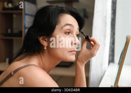 Una donna di 50 anni applica il mascara a casa, mostrando il suo volto naturale con rughe visibili, abbracciando la vera bellezza e la calma cura di sé davanti a una m. Foto Stock