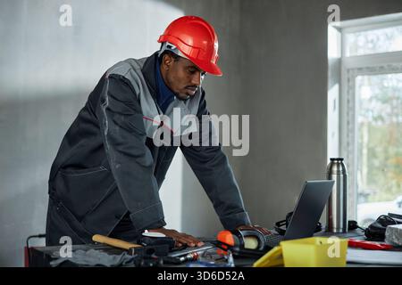 Black Man di mezza età che indossa un elmetto per lavorare su un laptop in interni Foto Stock