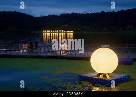 Una scultura di luna incandescente con figure di coniglio galleggia fuori dal passaggio pedonale del lago Tangeum mentre i ciclisti passano e il cartello CHUNGJU KOREA si riflette attraverso. Foto Stock