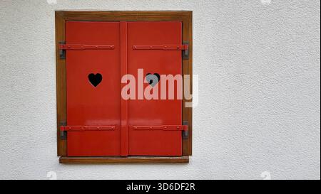 Vista ravvicinata delle persiane rosse rustiche a forma di cuore, incastonate all'interno di una cornice di legno contro una casa bianca pulita, Swiss Chalet Foto Stock