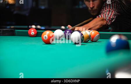 Palline da biliardo colorate sparse su un tavolo di feltro verde, mentre un uomo concentrato con una camicia a quadri punta il suo bastoncino sullo sfondo sfocato. Foto Stock