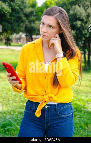 Donna pensiva con camicia gialla che tiene in mano lo smartphone all'aperto pensando al parco Foto Stock