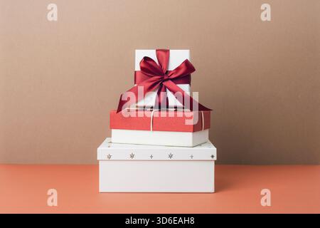 Scatole regalo impilate nei colori rosso e bianco con un fiocco in raso sulla parte superiore su sfondo beige, per una presentazione elegante e minimal. Foto Stock