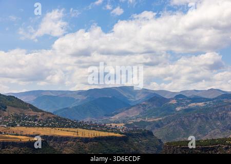 Ampio paesaggio caratterizzato da un villaggio a valle lungo aspre montagne, un cielo blu luminoso con nuvole bianche e colline a strati. Alaverdi, provincia di Lori Foto Stock