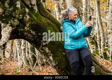 Una donna si appoggia su un albero coperto di muschio, scattando foto degli splendidi dintorni in una tranquilla foresta. La lussureggiante vegetazione mostra il caldo autunno Foto Stock