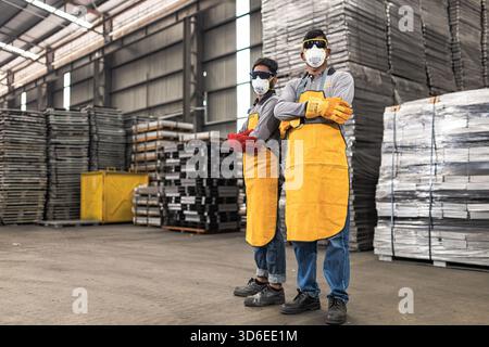 Due lavoratori industriali che indossano indumenti protettivi, tra cui maschere facciali, occhiali di sicurezza, camicie a maniche lunghe, grembiuli gialli e guanti per impieghi gravosi pose co Foto Stock