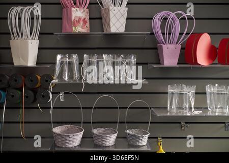 Cestelli per fiori vuoti assortiti, scatole a forma di cuore e vasi in plastica trasparente disposti su ripiani trasparenti contro pareti a righe, materiale per imballaggio floreale organizzato per l'esposizione Foto Stock