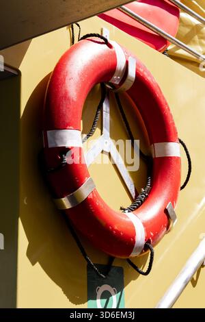 Un salvagente rosso brillante dotato di corde, attaccate a una parete gialla, che contrastano nettamente con la luce del sole. Foto Stock