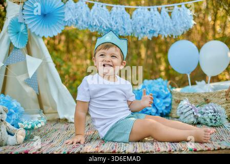 Un bambino allegro con una camicia bianca e pantaloncini a scacchi sorride, circondato da palloncini blu, fiori e decorazioni divertenti in un ambiente all'aperto, aggiungi Foto Stock