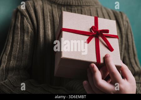Una foto ravvicinata che mostra le mani di una persona che tiene in mano una confezione regalo ben confezionata con un vivace nastro rosso sullo sfondo color ottanio. Foto Stock