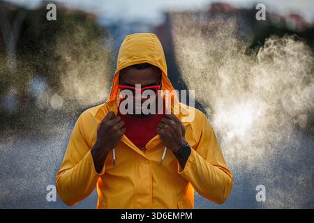 Uomo atletico con impermeabile giallo brillante e occhiali da sole rossi regola il cappuccio mentre la nebbia gli avvolge. Foto Stock