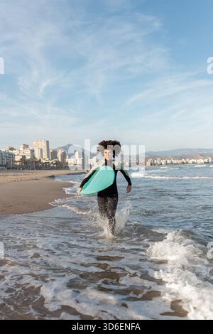 Giovane surfista donna premurosa in muta con tavola da surf che corre guardando via sulla riva bagnata dal mare ondeggiante Foto Stock