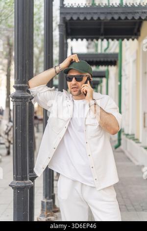 Un elegante uomo d'affari con una tuta bianca casual si appoggia a una classica lampada da strada mentre è impegnato in una conversazione con il suo smartphone in un'affascinante atmosfera Foto Stock