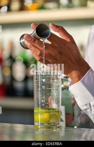 Un barista versa sapientemente del liquido in un alto bicchiere pieno di lime e menta, catturando l'atmosfera vivace di un classico mojito in un ambiente ben illuminato Foto Stock