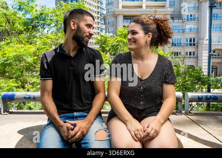 Un giovane uomo e una donna si siedono su una superficie di cemento in un parco cittadino, impegnandosi in una conversazione gioiosa con edifici moderni e lussureggiante vegetazione sul retro Foto Stock