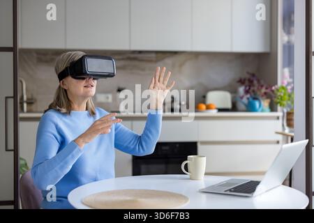 Donna anziana che indossa un visore vr e interagisce con lo spazio virtuale, seduto a un tavolo della cucina con un laptop, abbracciando la tecnologia moderna e la metaversa Foto Stock