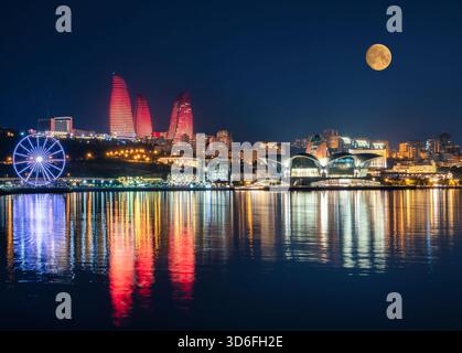 Vista notturna di Baku con le Torri delle fiamme, la ruota panoramica, il centro commerciale Deniz e la luna piena riflessa nel Mar Caspio. Foto Stock