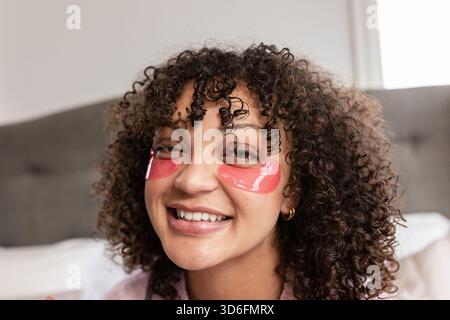 Donna con capelli ricci scuri che si sdraia sul letto in camera da letto applicando cerotti sotto gli occhi in idrogel rosa Foto Stock