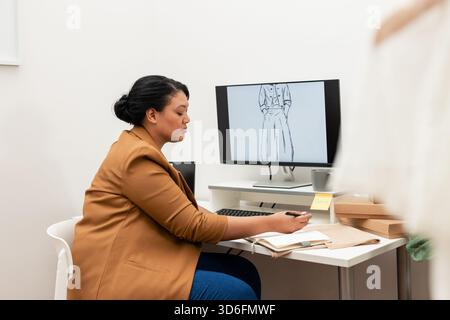 Donna afroamericana che studia disegno di moda sul monitor in casa studio con campioni di tessuto Foto Stock